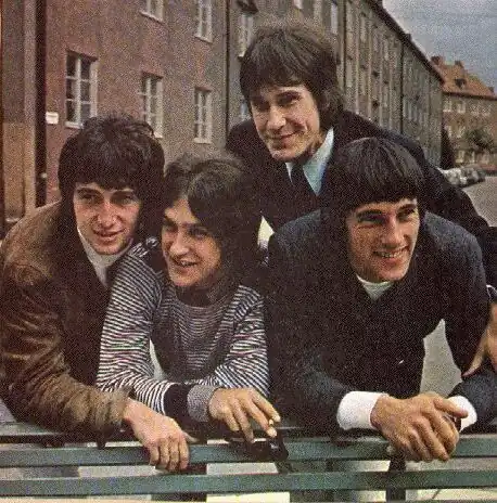 Four smiling men look around wearing moptops while standing behind a bench.