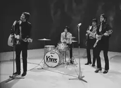 Four members of the band the Kinks stand onstage, during a television appearance. From left to right is Ray Davies, wearing a leather suit and long black pants. He is strumming an acoustic guitar and preparing to sing into a microphone placed in front of him. Next is Mick Avory, seated and playing the drums. He is wearing a brightly coloured long-sleeved shirt and dark pants. After him is bassist Pete Quaife, playing his instrument and wearing a set of clothes similar to Avory's. Last is Dave Davies, wearing what appears to be a leather suit with a short tie, black pants and high-heeled loafers. He plays an elaborate V-shaped electric guitar and is standing behind a microphone.