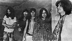 Five smiling men in a row, diagonal to the camera angle. The man on the left (farthest to the back) has very long hair and a full beard; he wears a white T-shirt and tie-dyed pants. Next to him, Dave Davies, also with very long hair, wears reflective sunglasses, a black short-sleeved shirt and jeans. In the middle, Mick Avory wears an unbuttoned leather vest and white pants. The man to his right wears a heavy, probably brown leather jacket with a design that is possibly Native American. On the far right, in front, Ray Davies wears a giant paisley kerchief knotted like a tie over a white jacket.